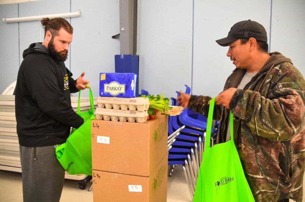 Two men stand in front of a table and fill shopping bags with food items. Cycles of poverty article.