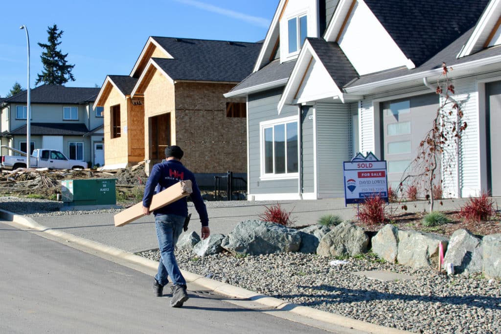 A worker carries building materials past a house with a “sold” sign.
